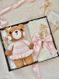 Crocheted teddy bear in a pink dress with a white blanket and dried flowers in a box.
