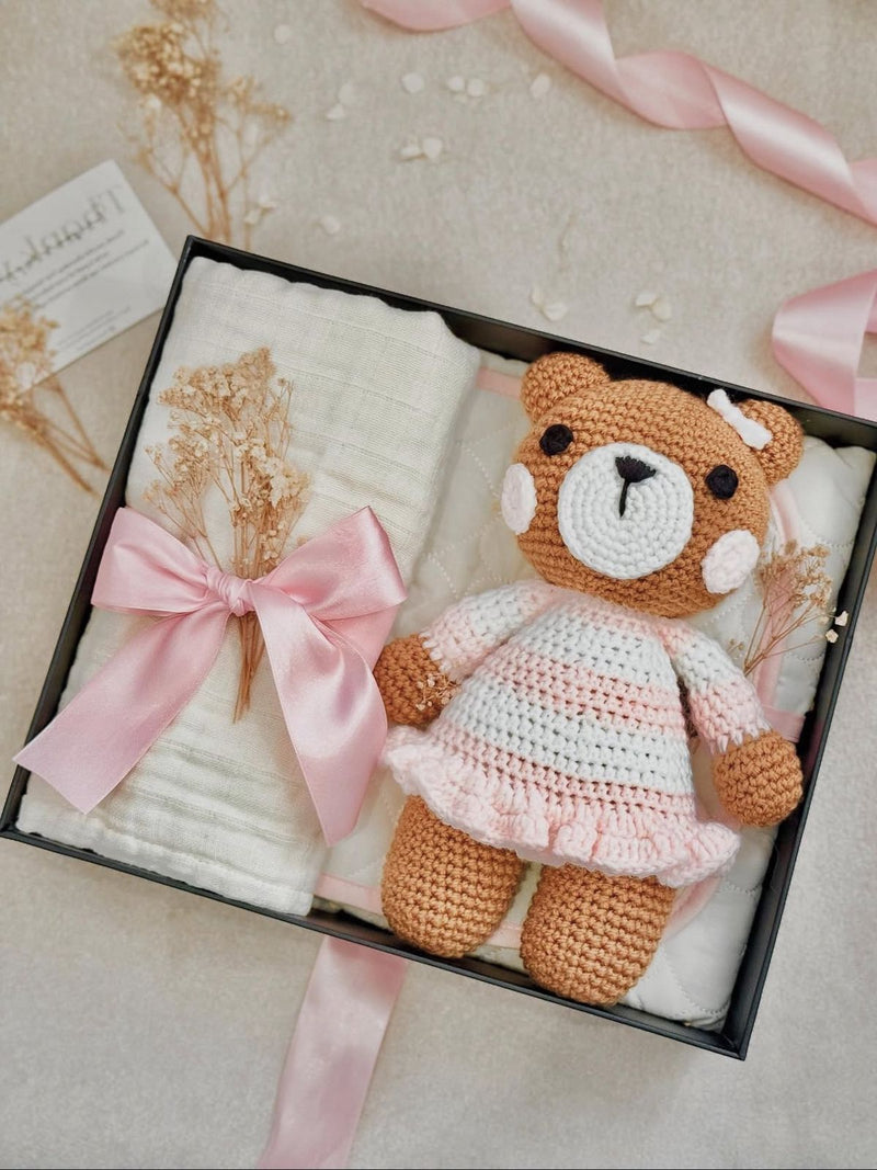 Crocheted teddy bear in a box with a pink bow and baby blanket on a light background