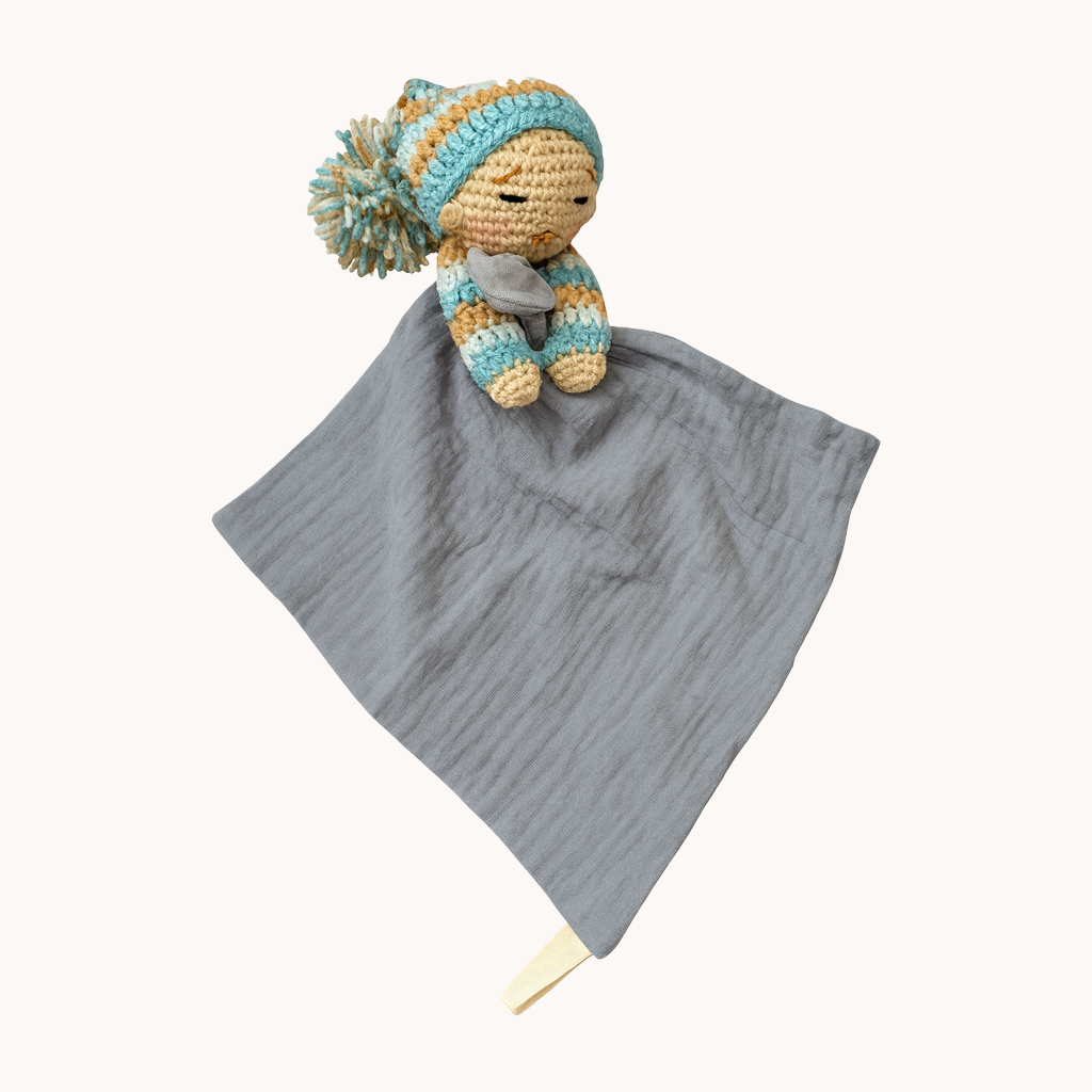 Crocheted baby comfort toy with pom-pom hat on a gray blanket against a white background